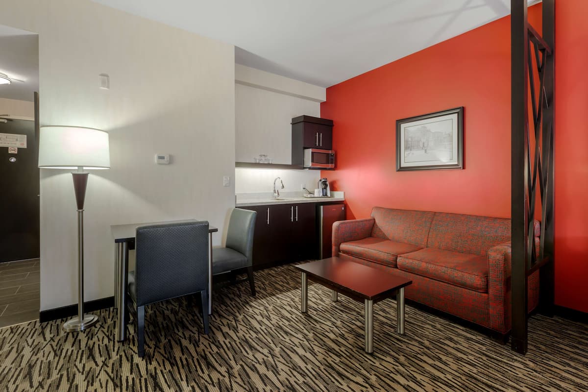 Una habitación de hotel compacta con kitchenette, sofá, mesa y lámpara de pie.