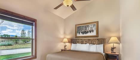 A bedroom with a large bed, two bedside lamps, a ceiling fan, a window with a view of a landscape, and a framed picture on the wall.