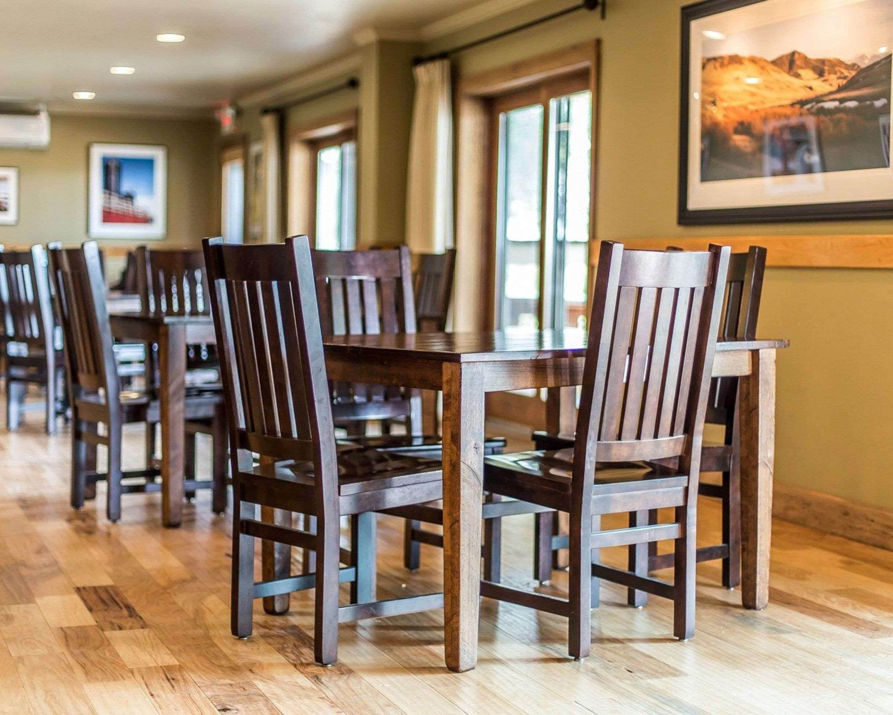 A dining area with wooden tables and chairs, large windows, and framed pictures on the walls.