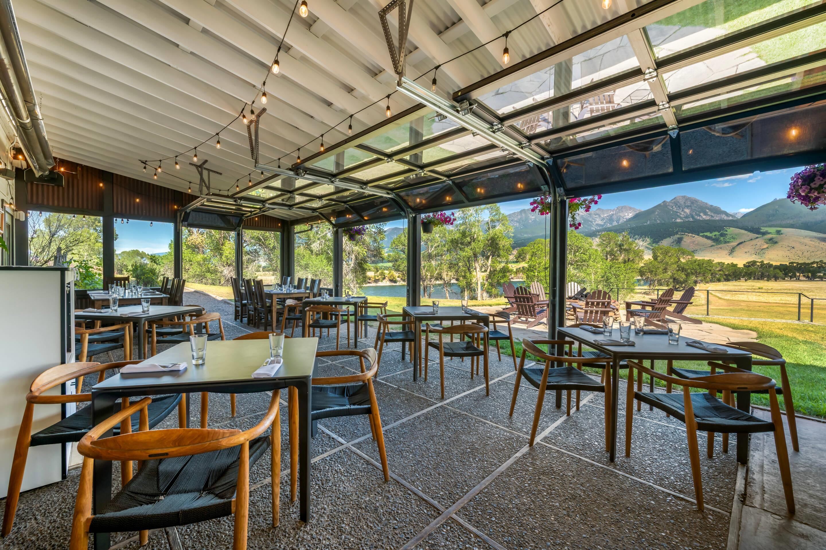 An outdoor dining area with tables and chairs, a glass ceiling, and a view of a mountainous landscape.