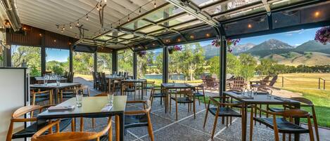 An outdoor dining area with tables and chairs, a glass ceiling, and a view of a mountainous landscape.