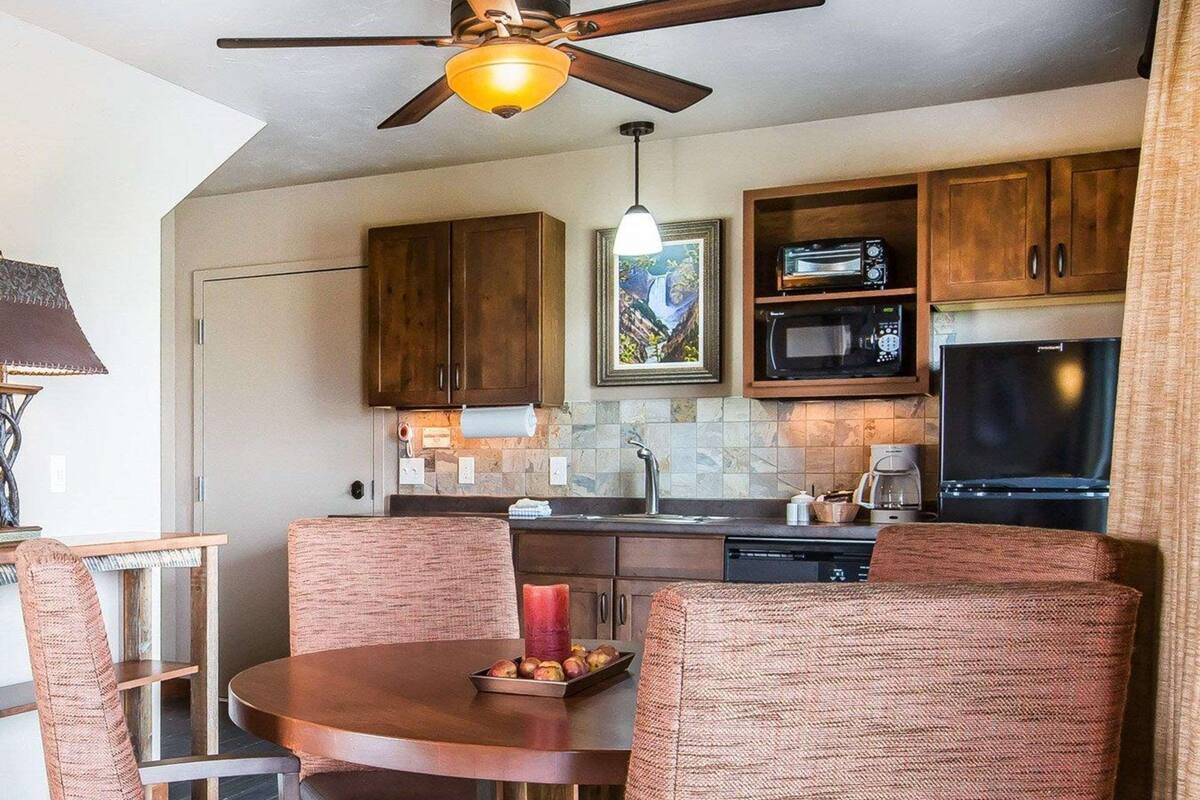 A compact kitchen with wooden cabinets, a microwave, a coffee maker, and a refrigerator. There is a round table with chairs and a ceiling fan above.
