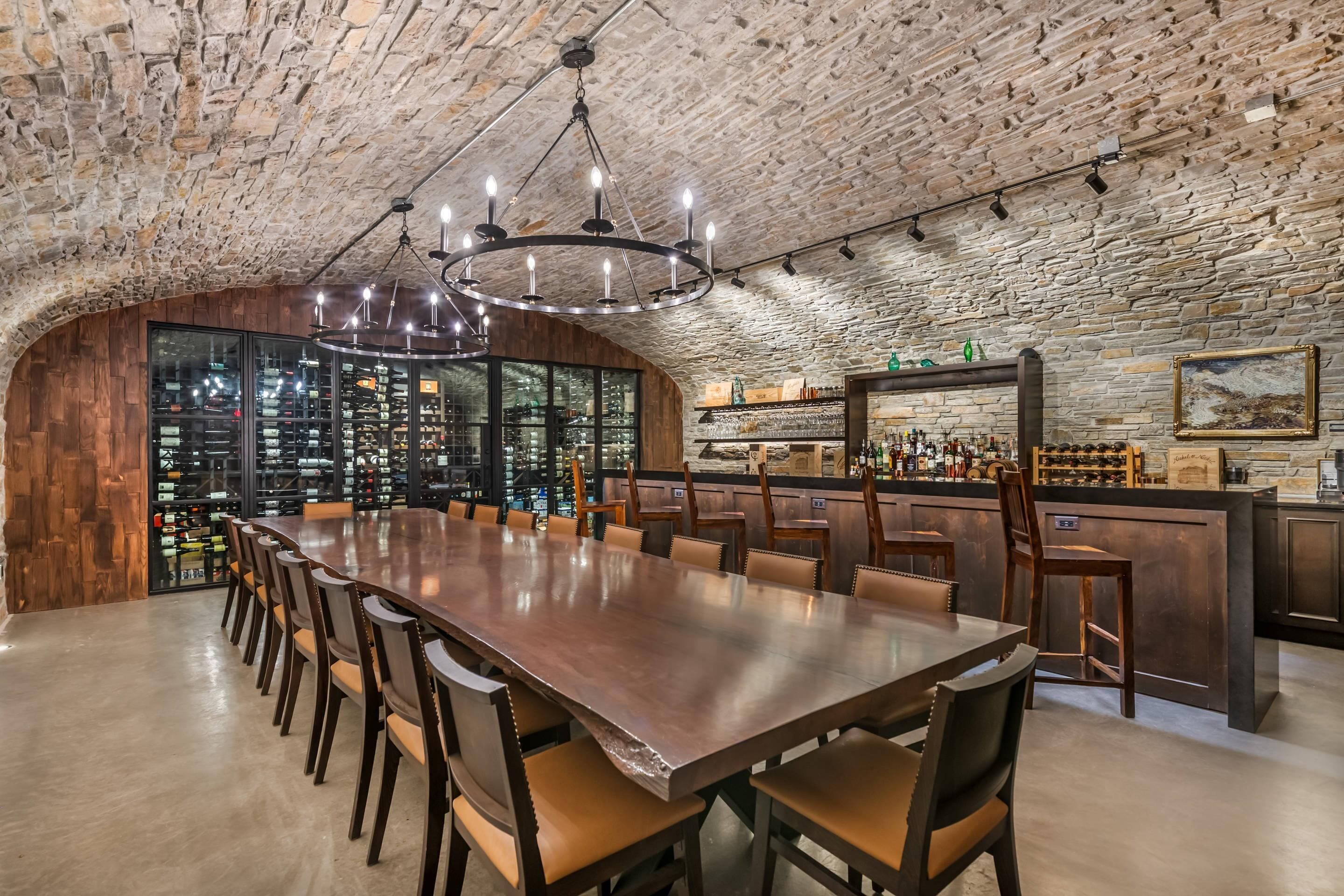 A spacious bar area with a long wooden table and chairs, a stone wall backdrop, and a variety of bottles and glasses on shelves.