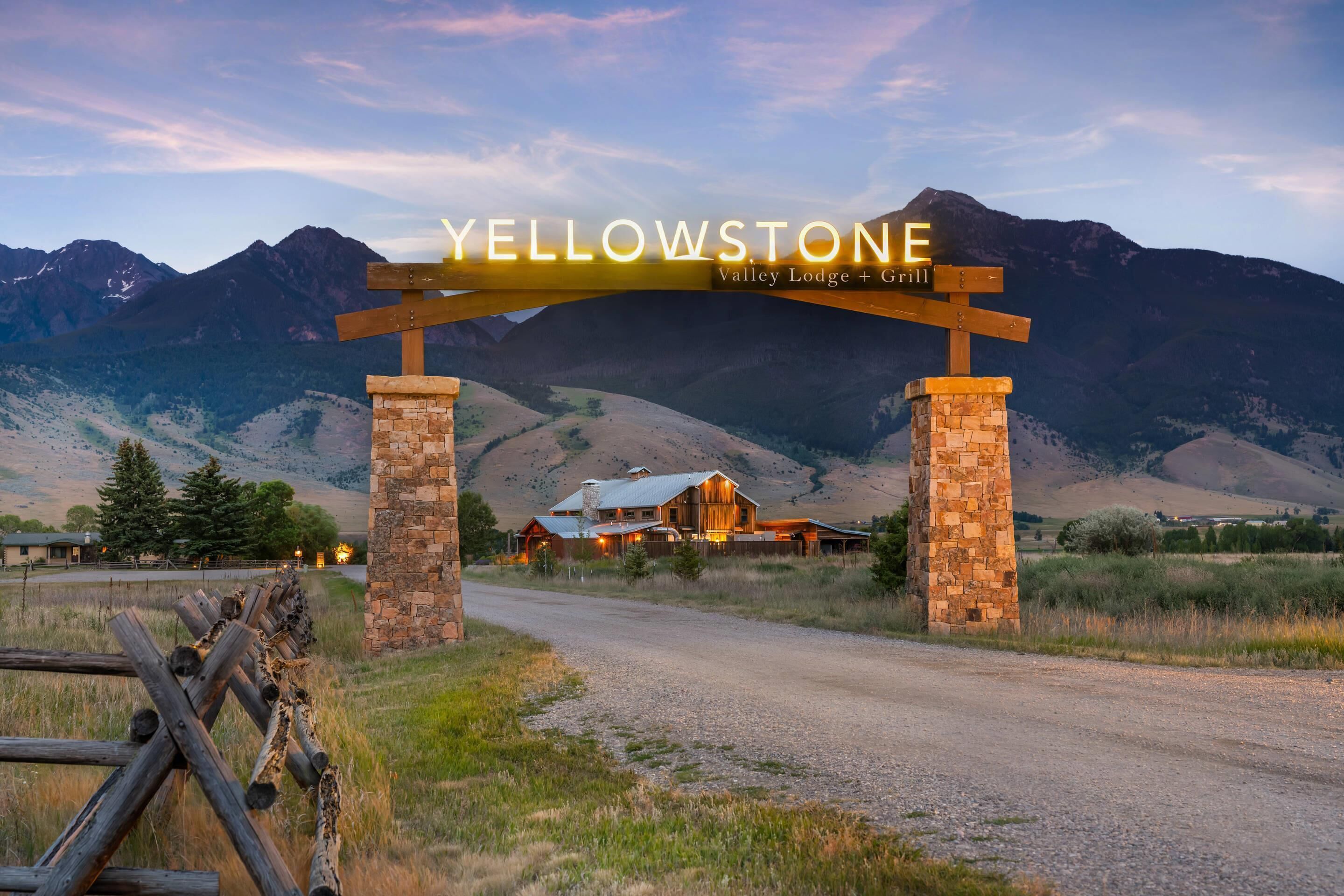 A wooden archway with the text 'YELLOWSTONE Valley Lodge + Grill' over a gravel road leading to a lodge with a metal roof, surrounded by mountains.
