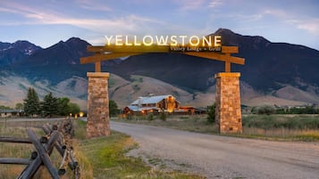 A wooden archway with the text 'YELLOWSTONE Valley Lodge + Grill' over a gravel road leading to a lodge with a metal roof, surrounded by mountains.