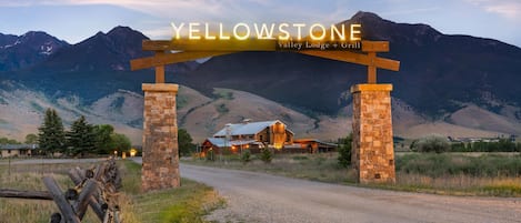 A wooden archway with the text 'YELLOWSTONE Valley Lodge + Grill' over a gravel road leading to a lodge with a metal roof, surrounded by mountains.