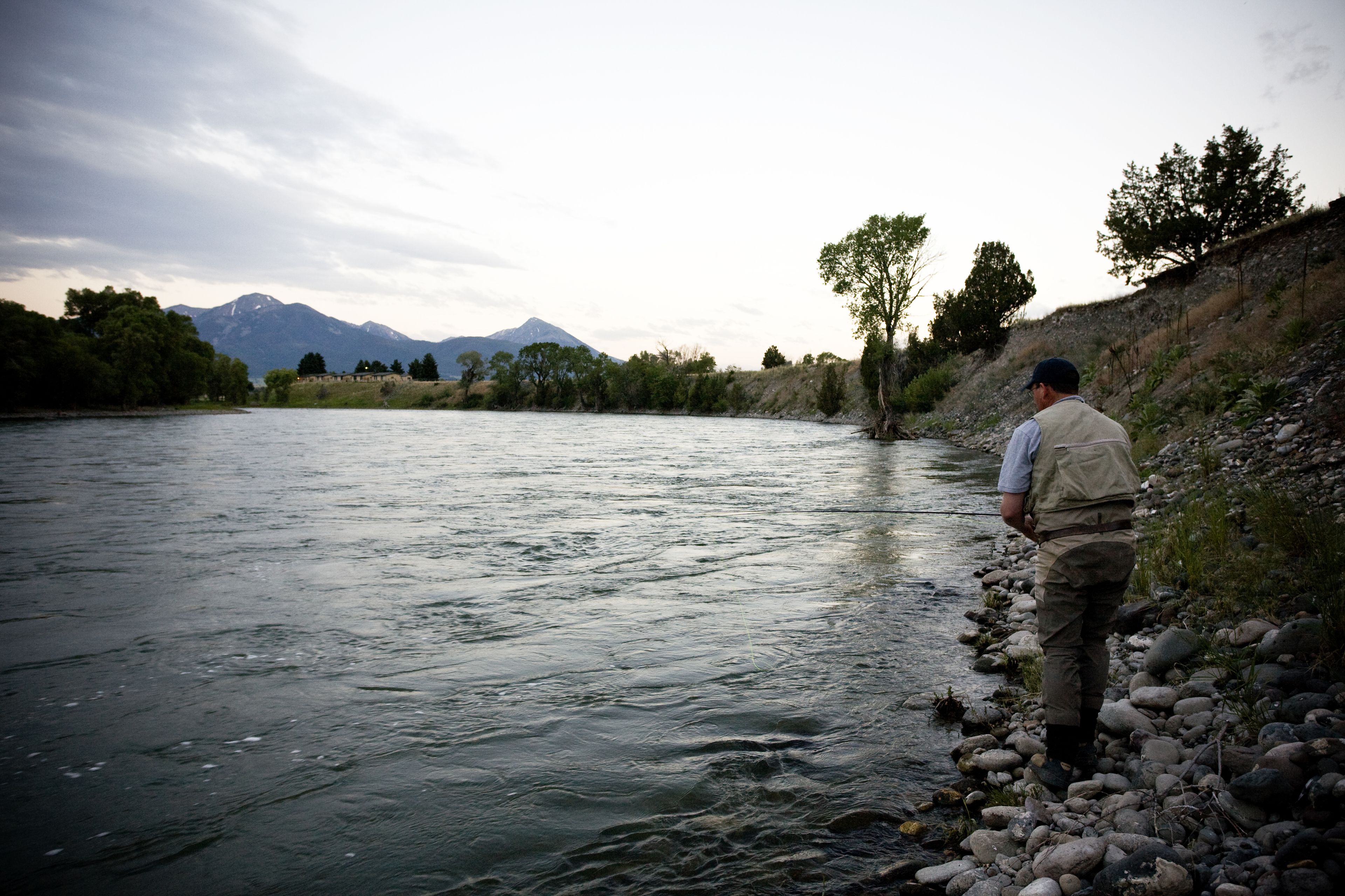 A man standing by a river with mountains in the background.