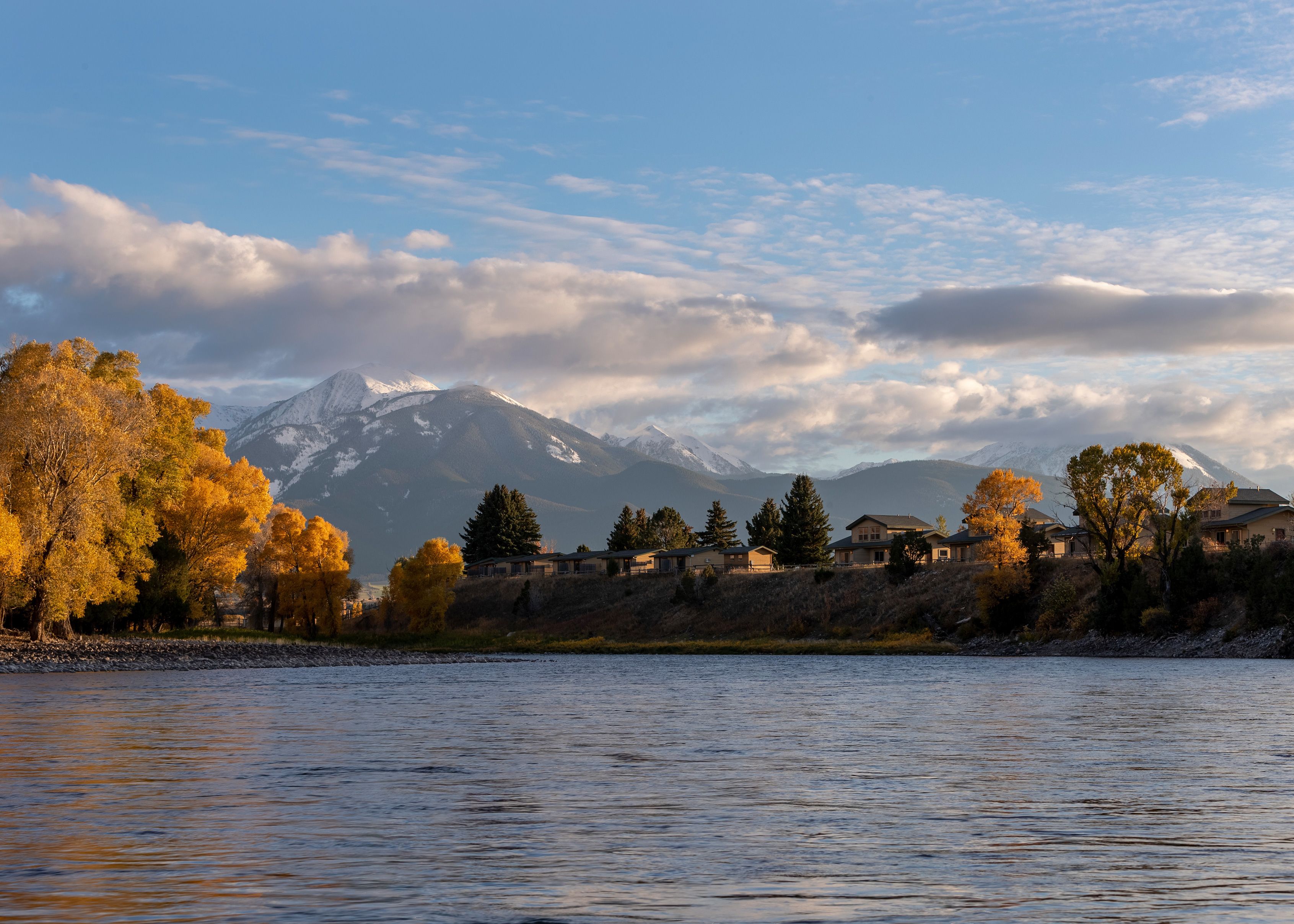 Une vue sur un lac, avec une chaîne de montagnes en arrière-plan, des arbres aux couleurs automnales et un petit village avec des maisons.
