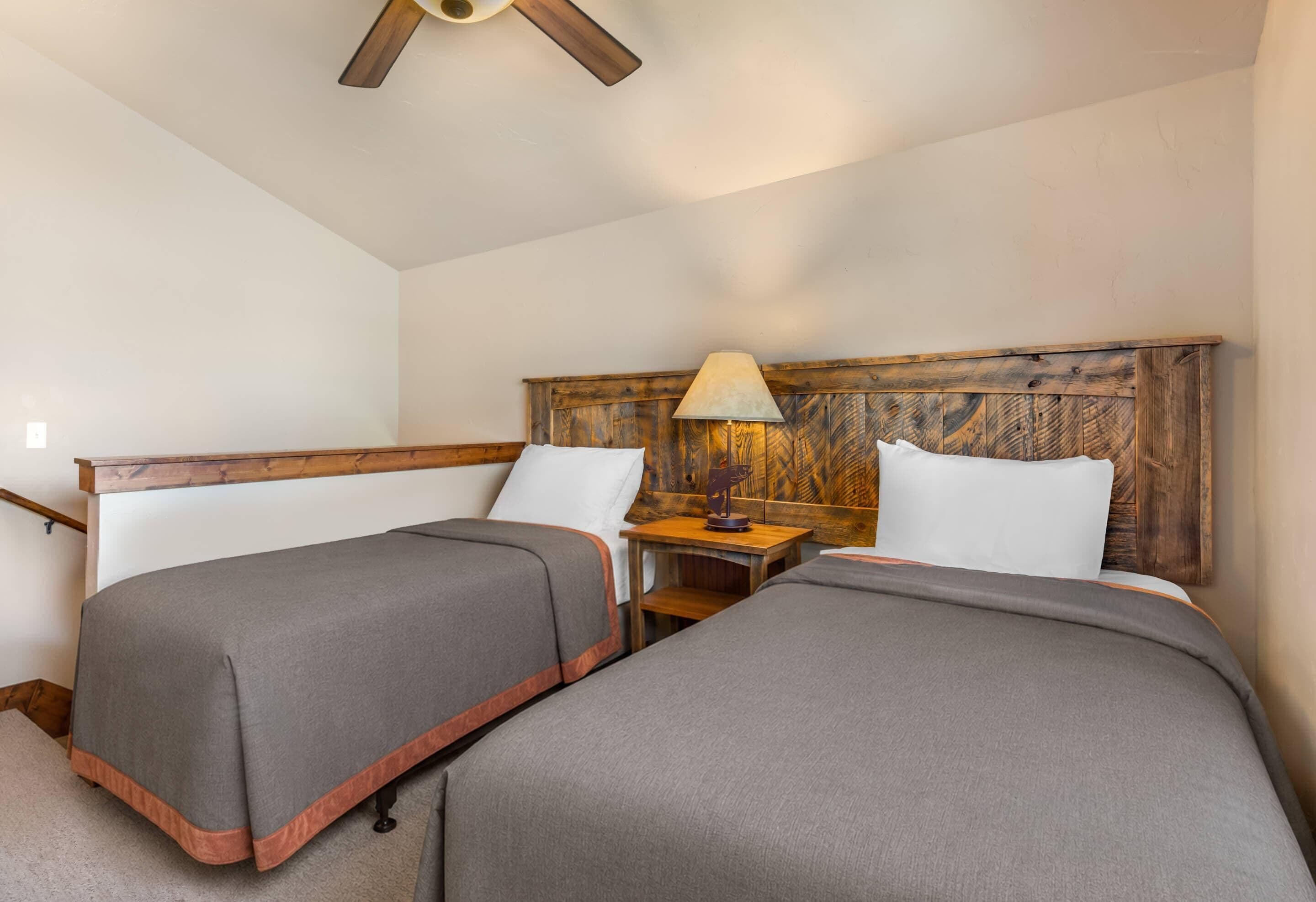 Habitación de hotel con dos camas, cabecera de madera, mesita de noche con lámpara y ventilador de techo.