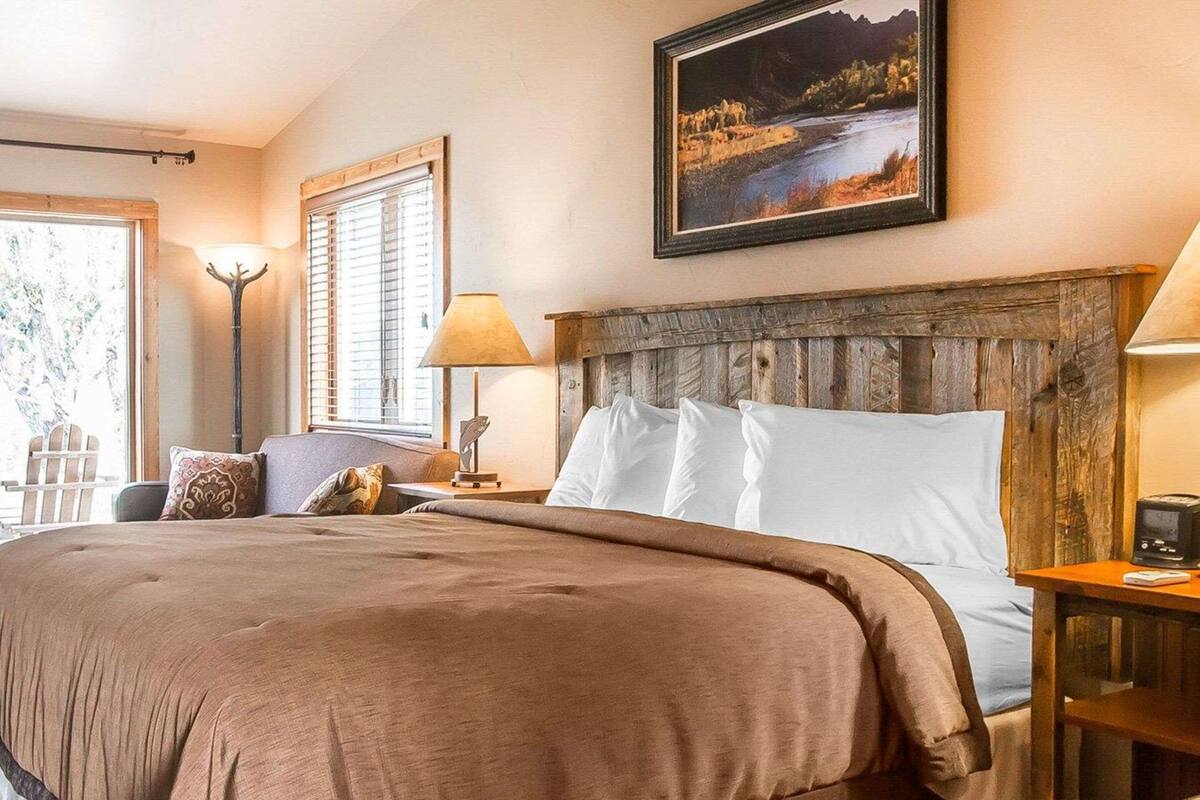 A hotel room with a large bed, a wooden headboard, a painting on the wall, and a window with blinds.