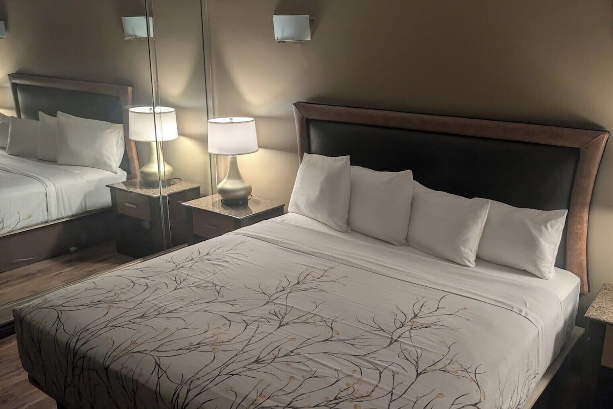 A hotel room with a large bed, two bedside lamps, a nightstand, a mirror, and a wooden headboard.