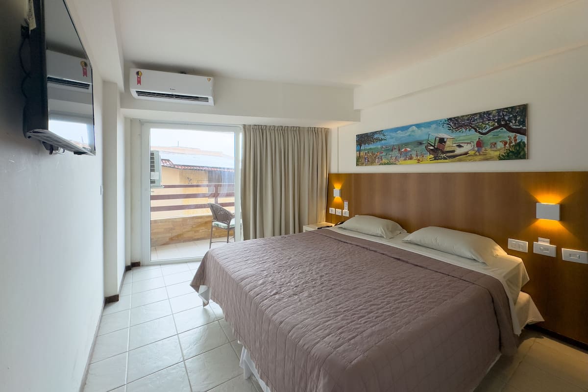 A hotel room with a bed, a painting on the wall, a window with curtains, and an air conditioning unit.