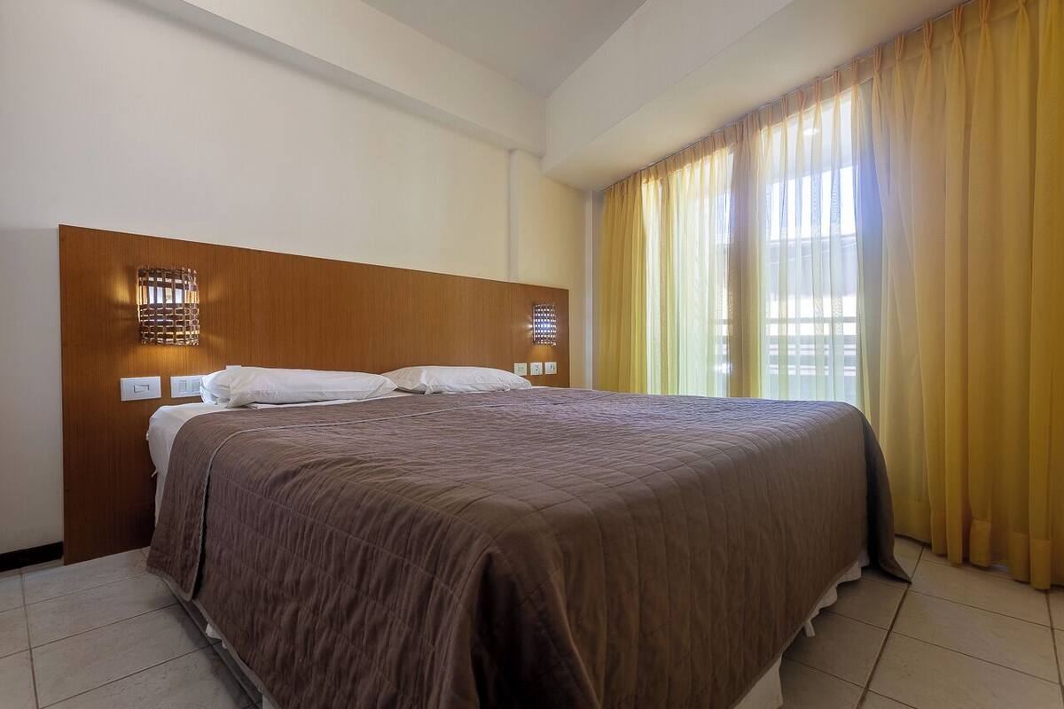 A hotel room with a large bed, wooden headboard, two wall-mounted light fixtures, and a window with yellow curtains.