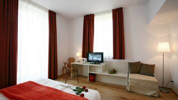 Una camera d'albergo con un letto, una scrivania con un computer, una sedia, una TV e un comodino con una lampada.