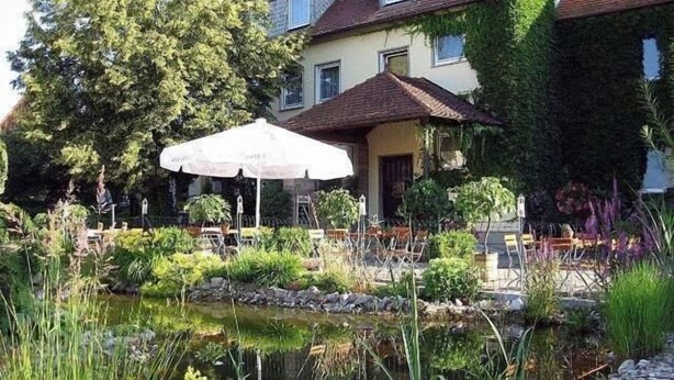 A garden with a pond, outdoor seating, and a building with a tiled roof. - Landgasthof Wellmann (Markt Taschendorf)
