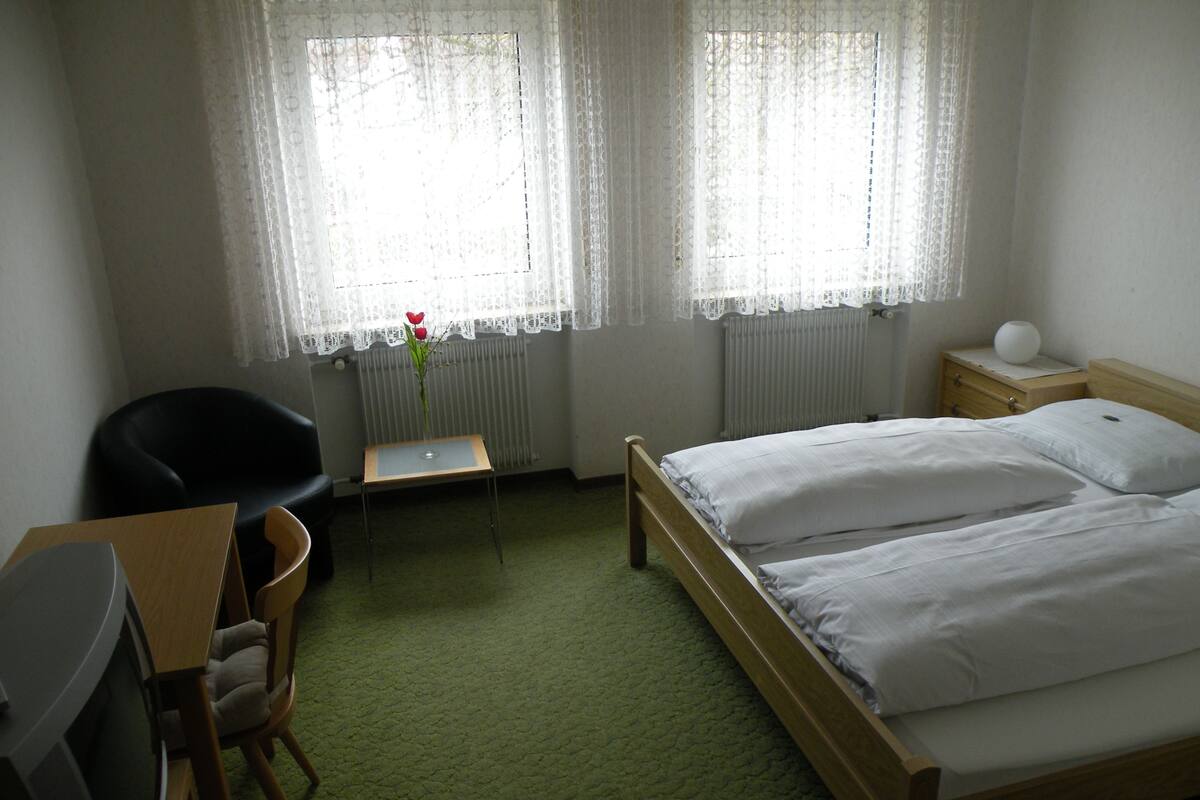 Une chambre d’hôtel avec un lit, un bureau et une chaise, un radiateur et une fenêtre avec des rideaux.
