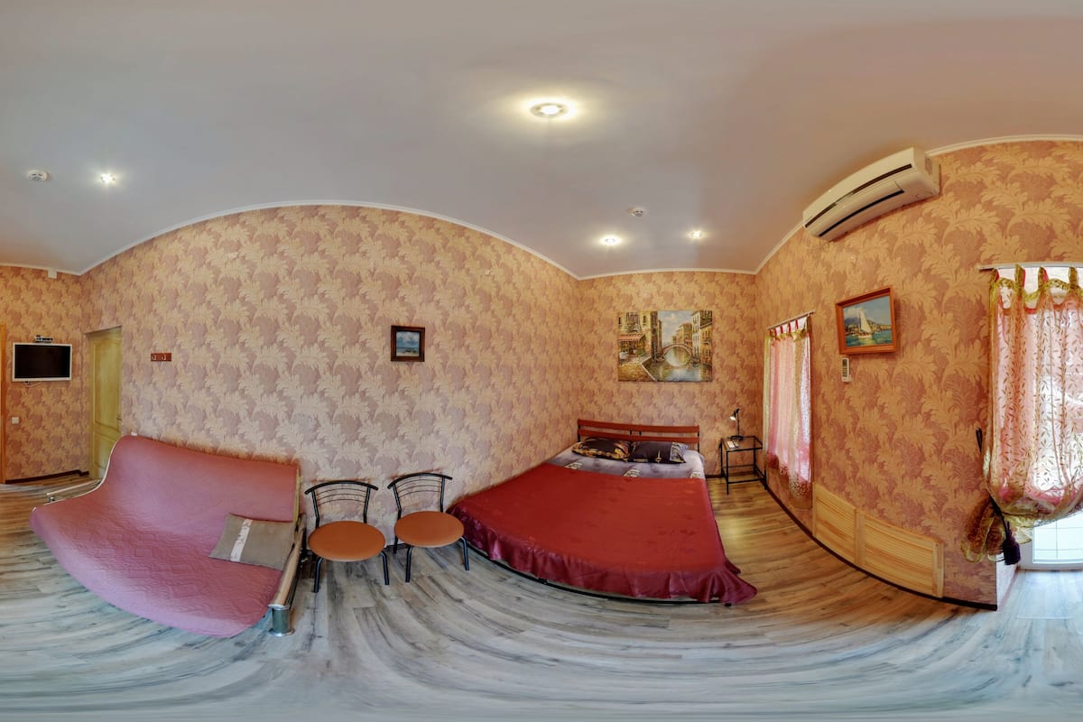 A panoramic view of a hotel room with a curved ceiling, patterned wallpaper, a bed with a red bedspread, a pink sofa, two chairs, a small table, and an air conditioning unit.