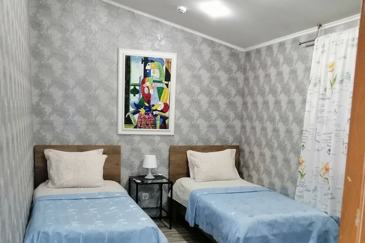 A room with two beds, a small table, a lamp, and a painting on the wall.