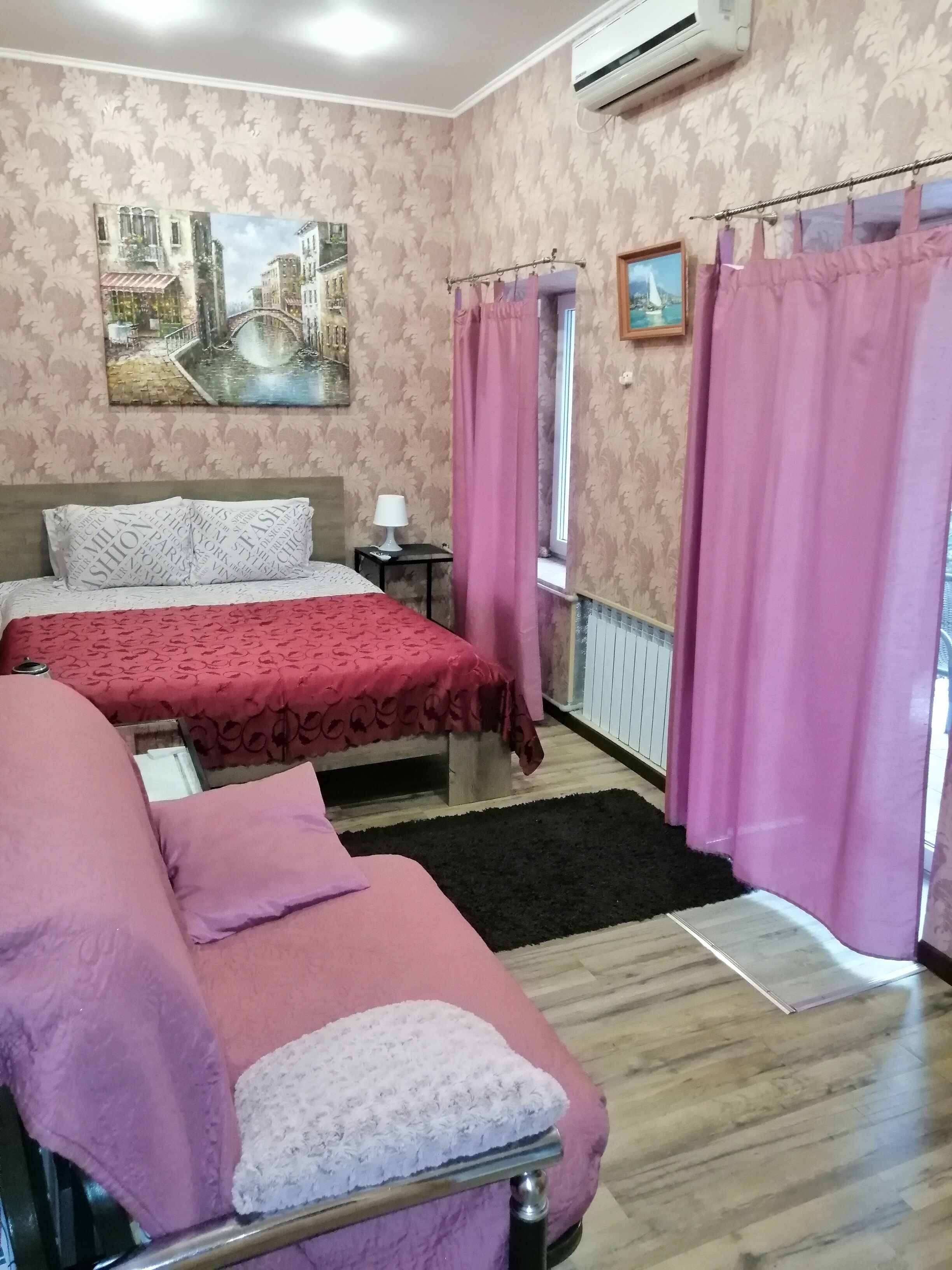 Een hotelkamer met twee bedden, een raam met roze gordijnen, een schilderij aan de muur en een airconditioner.