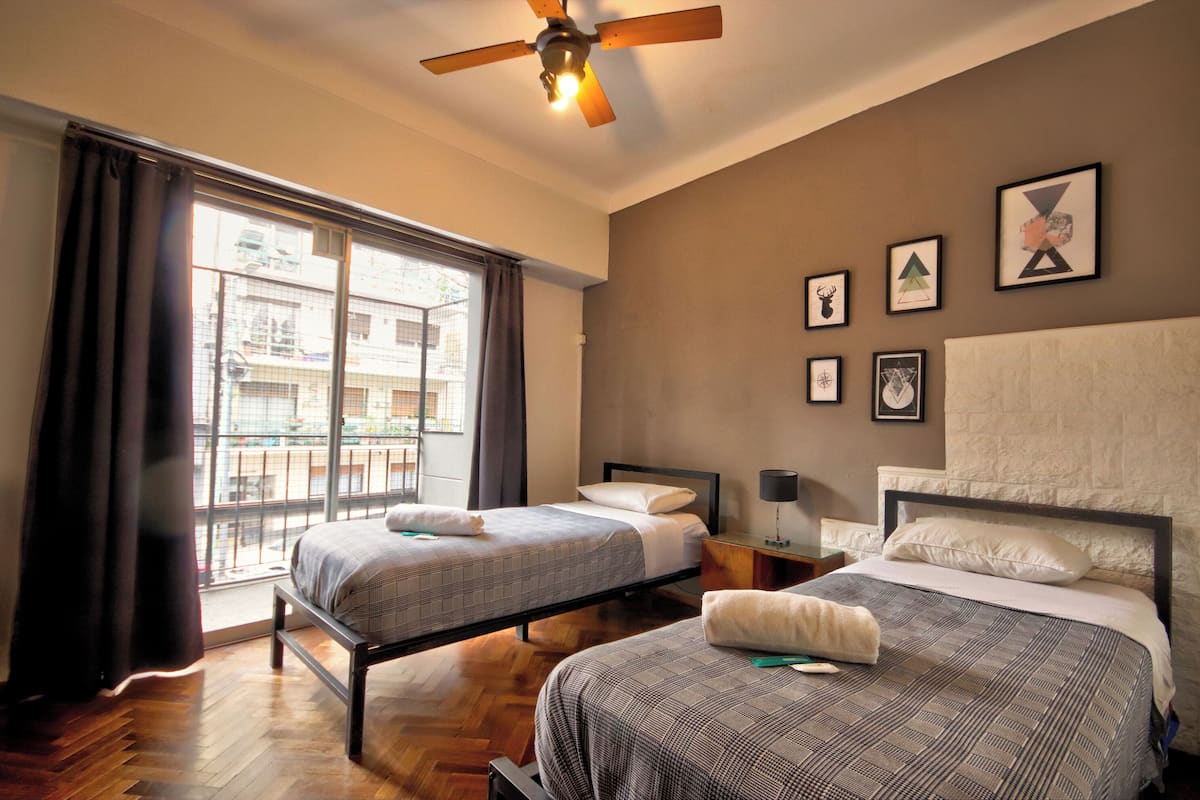 A hotel room with two beds, a ceiling fan, a balcony with a view of buildings, and several framed pictures on the wall.