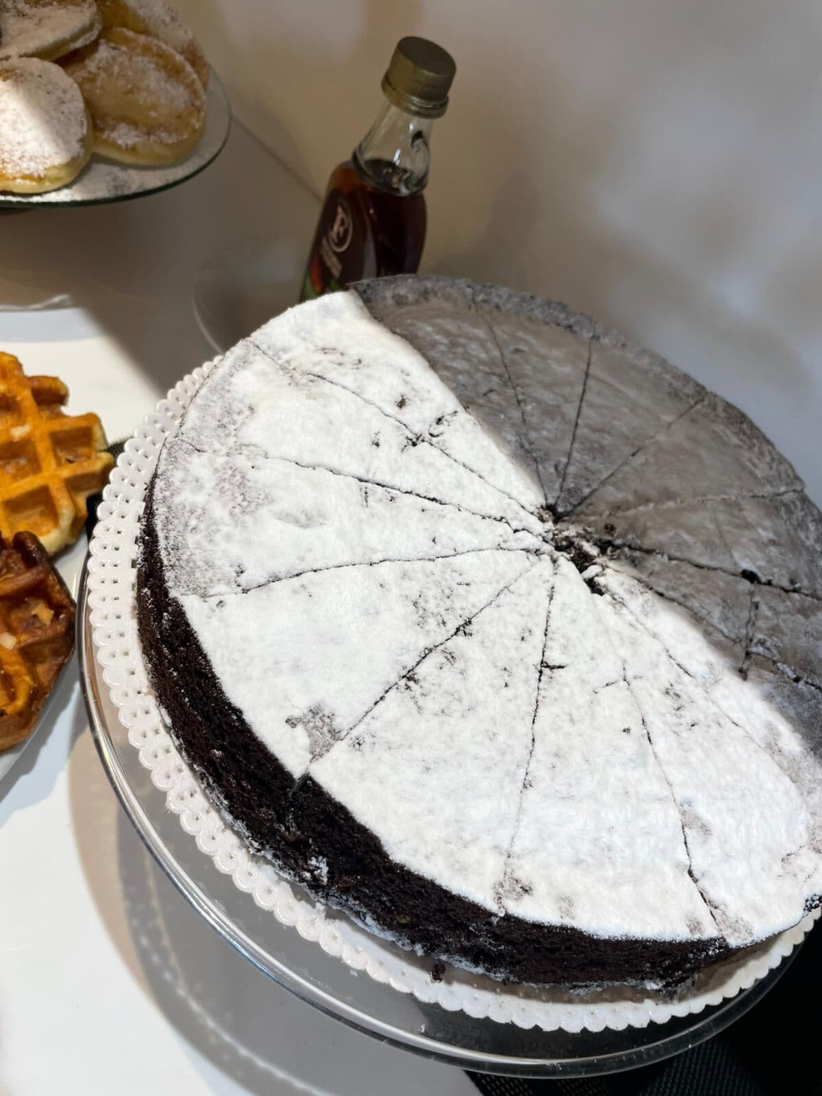 A round cake with a white frosting and a dusting of powdered sugar on top, placed on a silver cake stand.