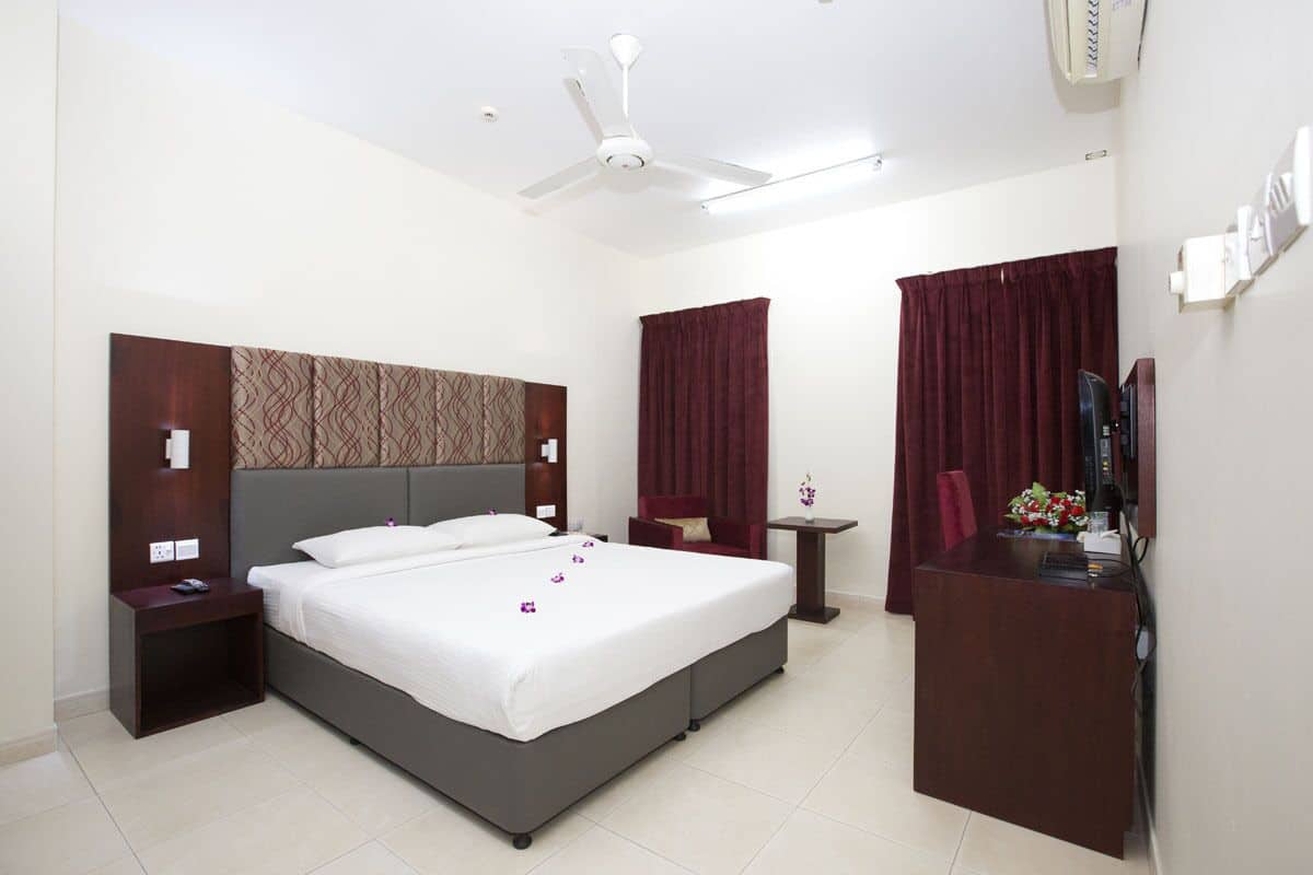 A hotel room with a large bed, a desk, a chair, a small table, a ceiling fan, and a TV.