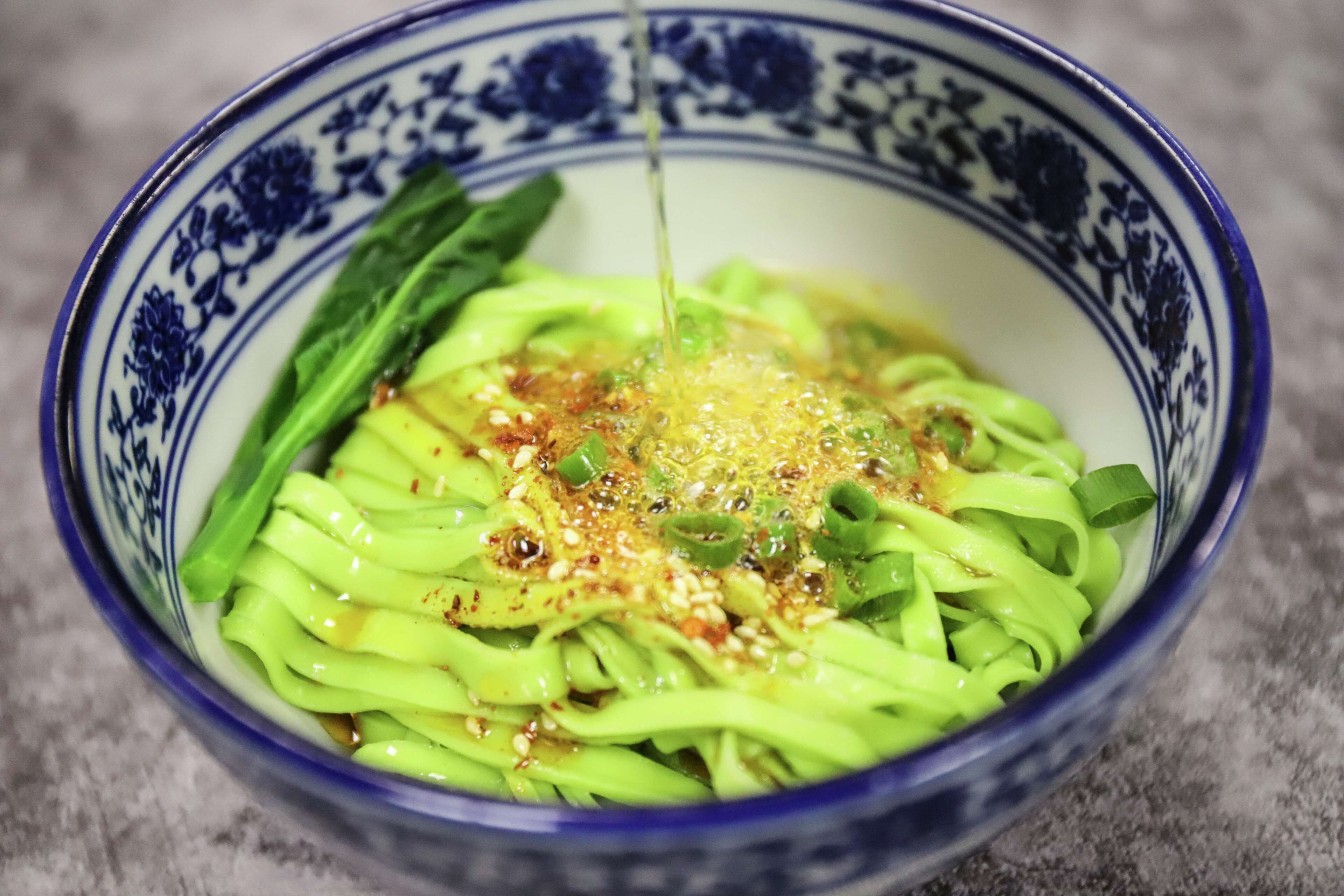 A bowl of green noodles with a clear liquid being poured over them, garnished with green onions and sesame seeds.