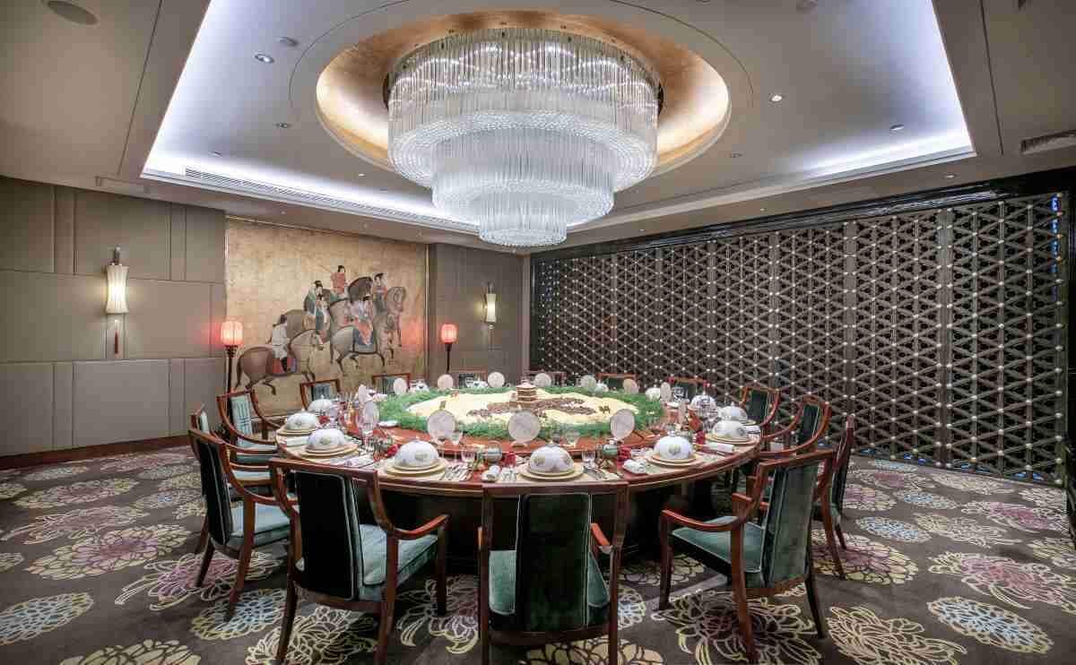A round dining table set for a meal with a chandelier above, surrounded by chairs with green upholstery.