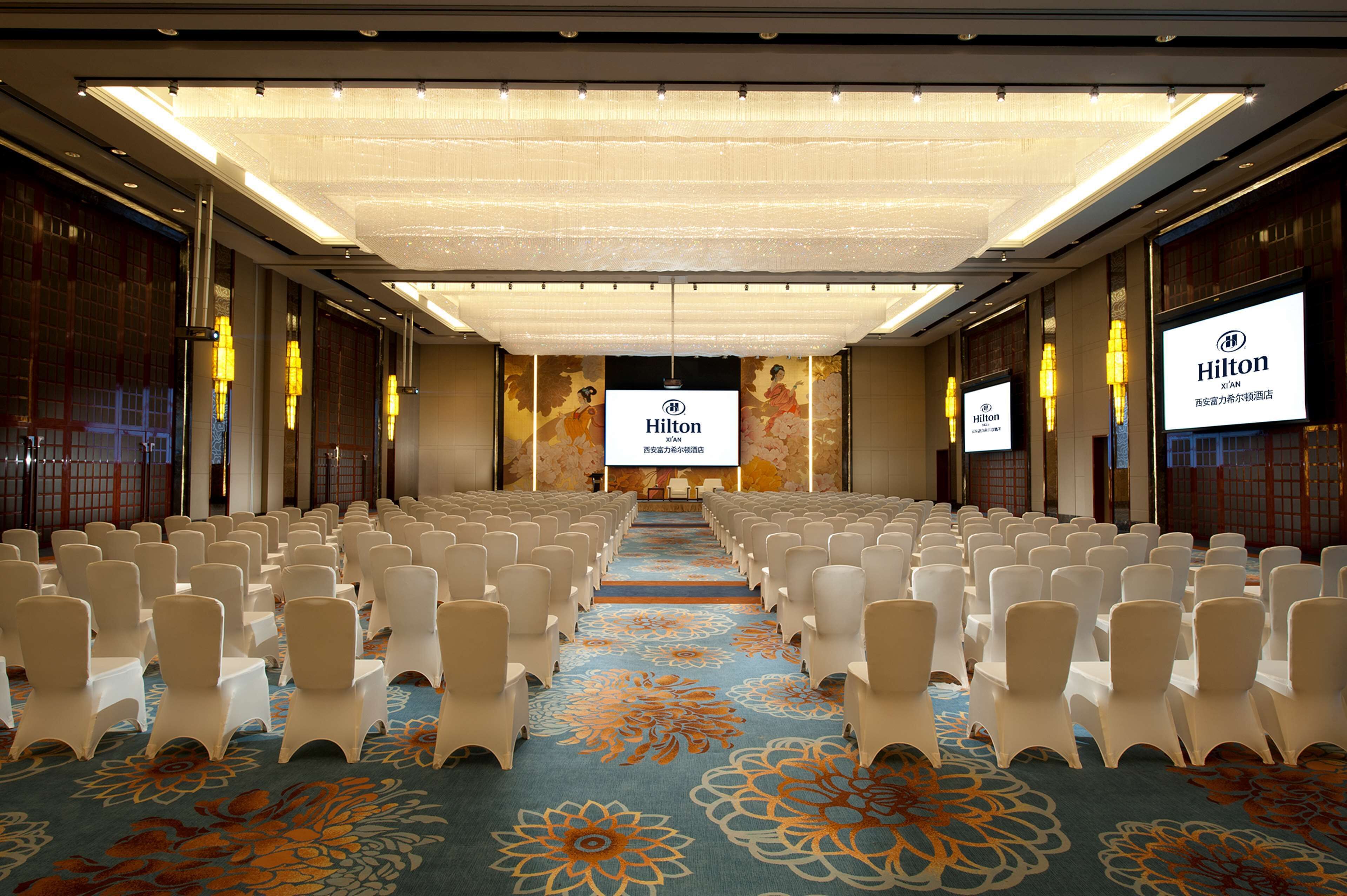 A conference room with rows of white chairs, a carpet with floral patterns, and a large screen displaying the Hilton logo.