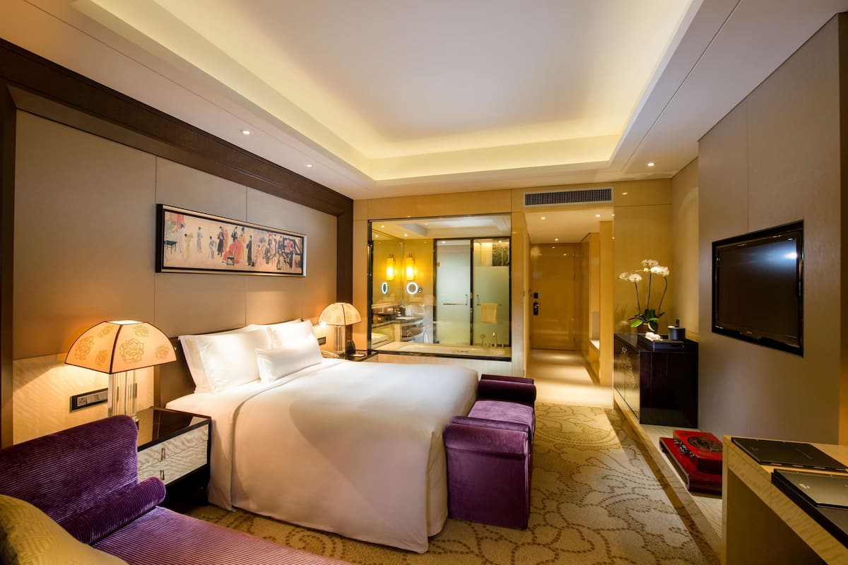 A hotel room with a large bed, two bedside tables with lamps, a TV, a sofa, and a small table with a red tray.