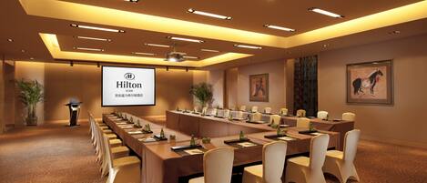 A conference room with a long table, chairs, a projector screen, and a potted plant.