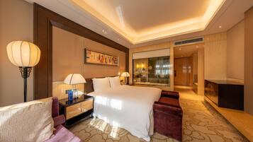A hotel room with a large bed, a sitting area with a sofa and armchair, a desk with a lamp, and a large window.