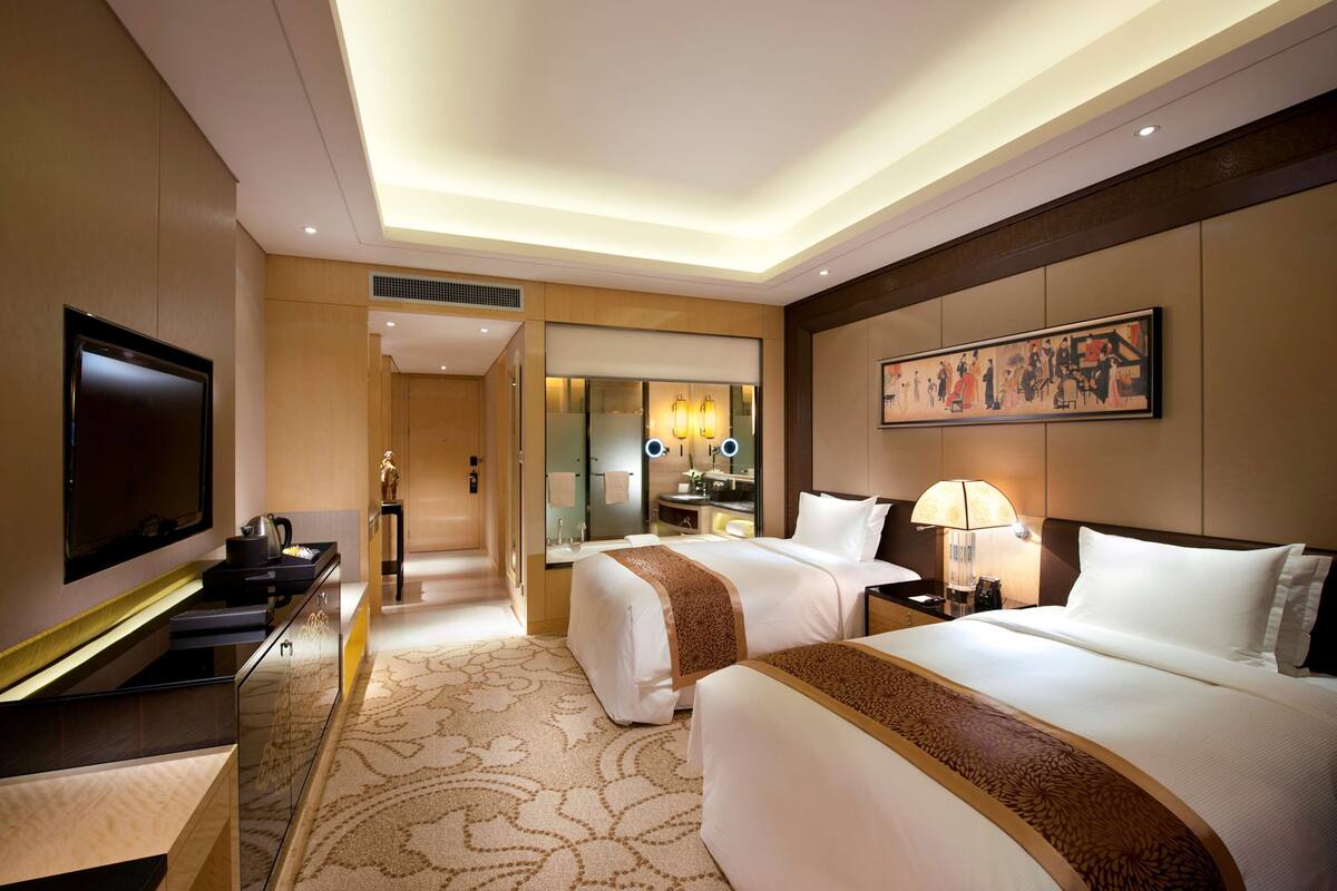A hotel room with two beds, a TV, a desk, and a bathroom with a mirror and lights.