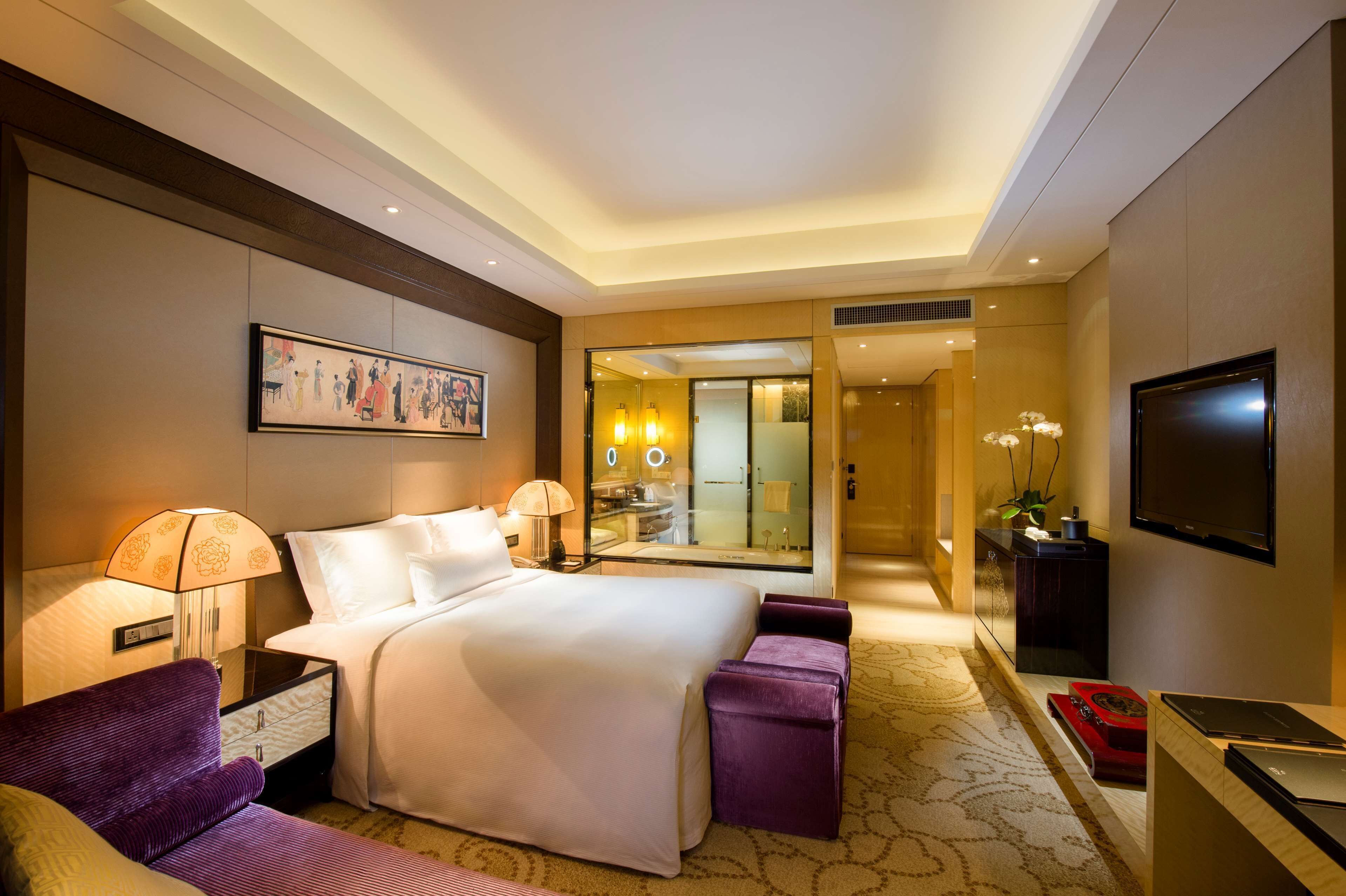 A hotel room with a large bed, a desk with a flat-screen TV, a sofa, a mirror, and a bathroom visible through an open door.