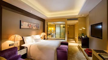 A hotel room with a large bed, a desk with a flat-screen TV, a sofa, a mirror, and a bathroom visible through an open door.