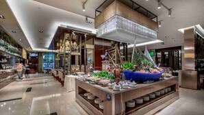 Un moderno vestíbulo de hotel con una zona de bufé central que presenta un recipiente azul grande lleno de comida, rodeado de platos y tazones.