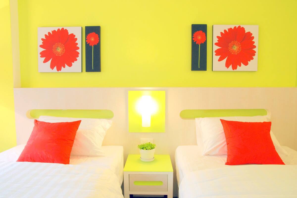 Een hotelkamer met twee bedden, elk voorzien van een rode kussen en witte beddengoed. Boven de bedden hangen twee bloemschilderijen. Een klein nachtkastje met een groene bovenkant en een witte lade staat tussen de bedden.