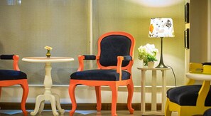 A hotel room with a seating area featuring two chairs with orange and black upholstery, a small round table with a vase of flowers, and a floor lamp with a patterned lampshade.