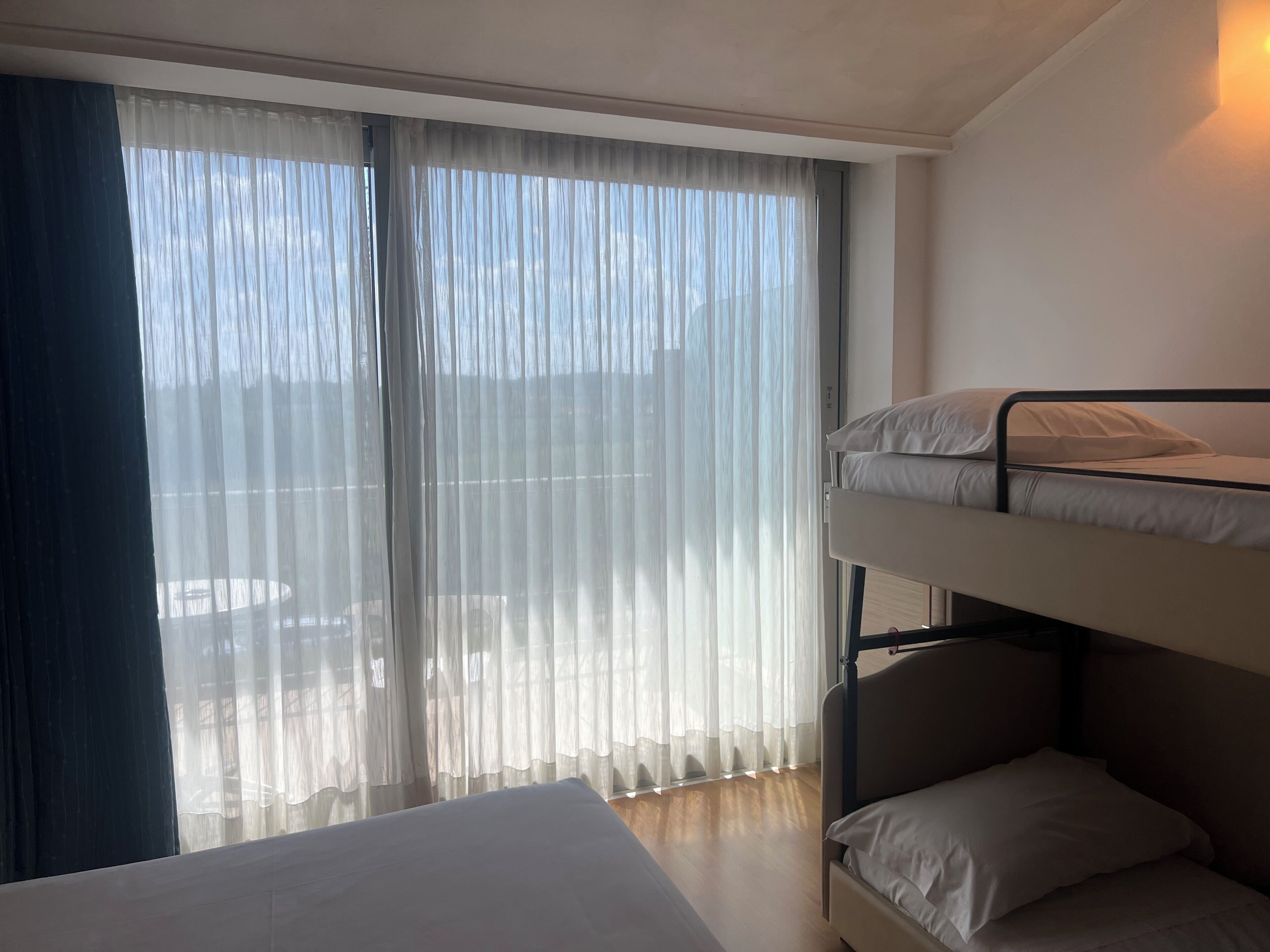 A room with a bunk bed, a large window with sheer curtains, and a bed with white bedding.