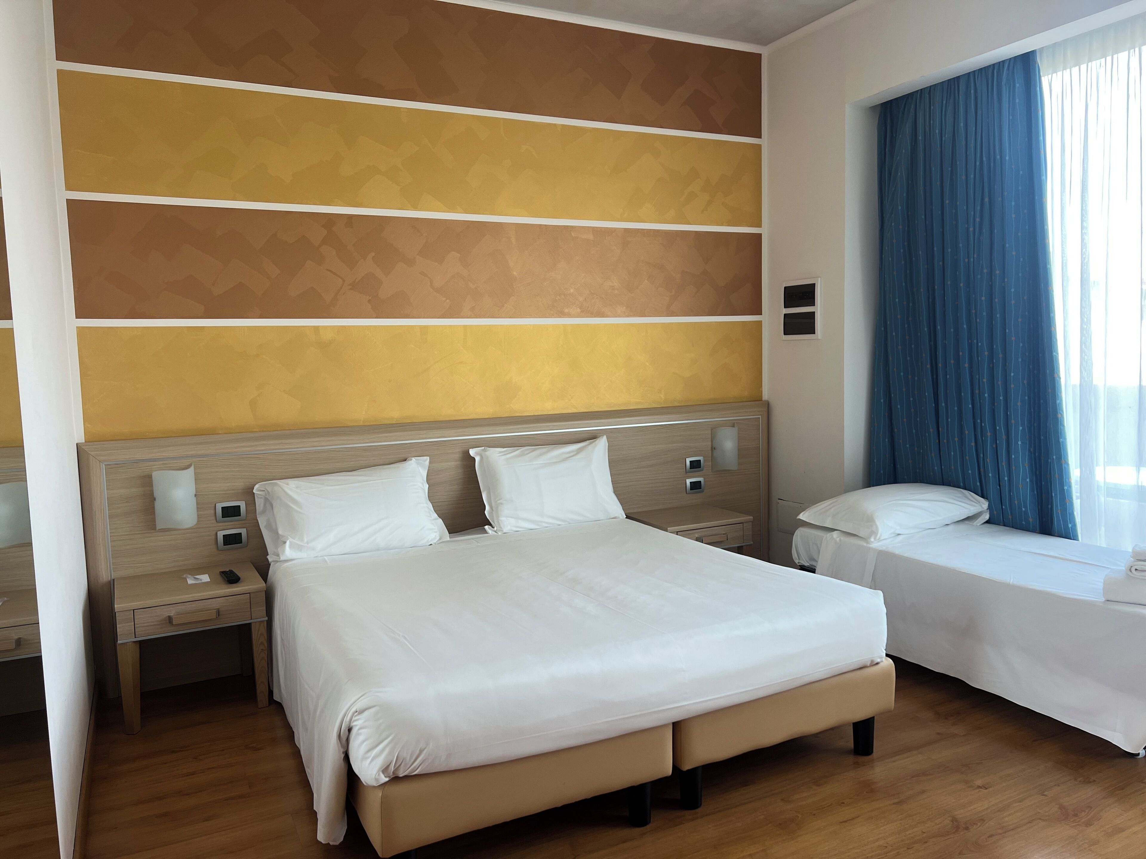 Une chambre d’hôtel avec un grand lit, un lit plus petit, une table de chevet et une fenêtre avec des rideaux.