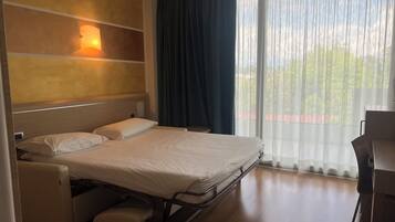 A hotel room with a bed, a bedside table, a chair, a wooden headboard, a wall-mounted light, and a view through sheer curtains.