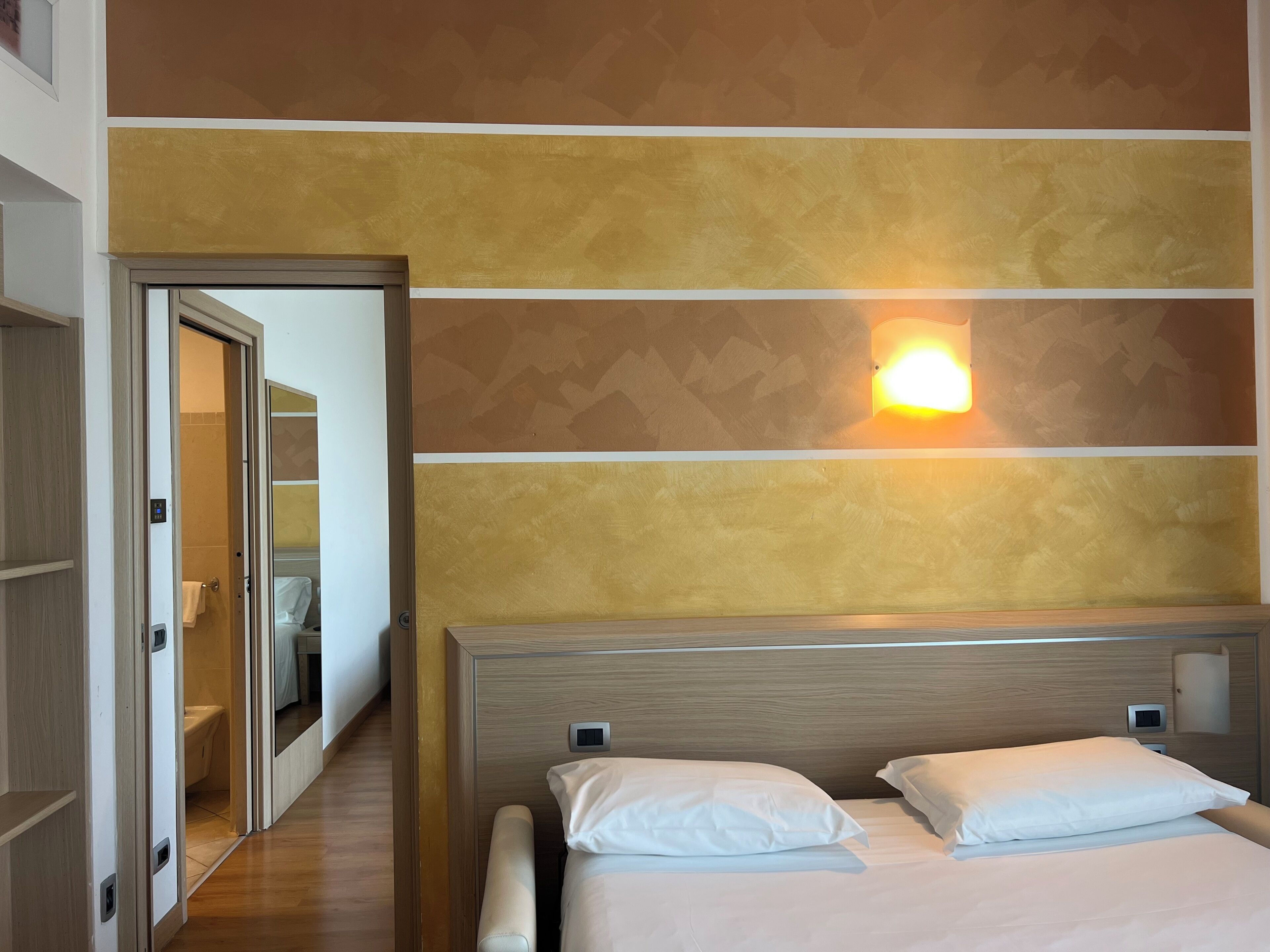 Une chambre d’hôtel avec une tête de lit en bois, un lit recouvert de draps blancs, une lampe fixée au mur et une porte donnant accès à la salle de bain.