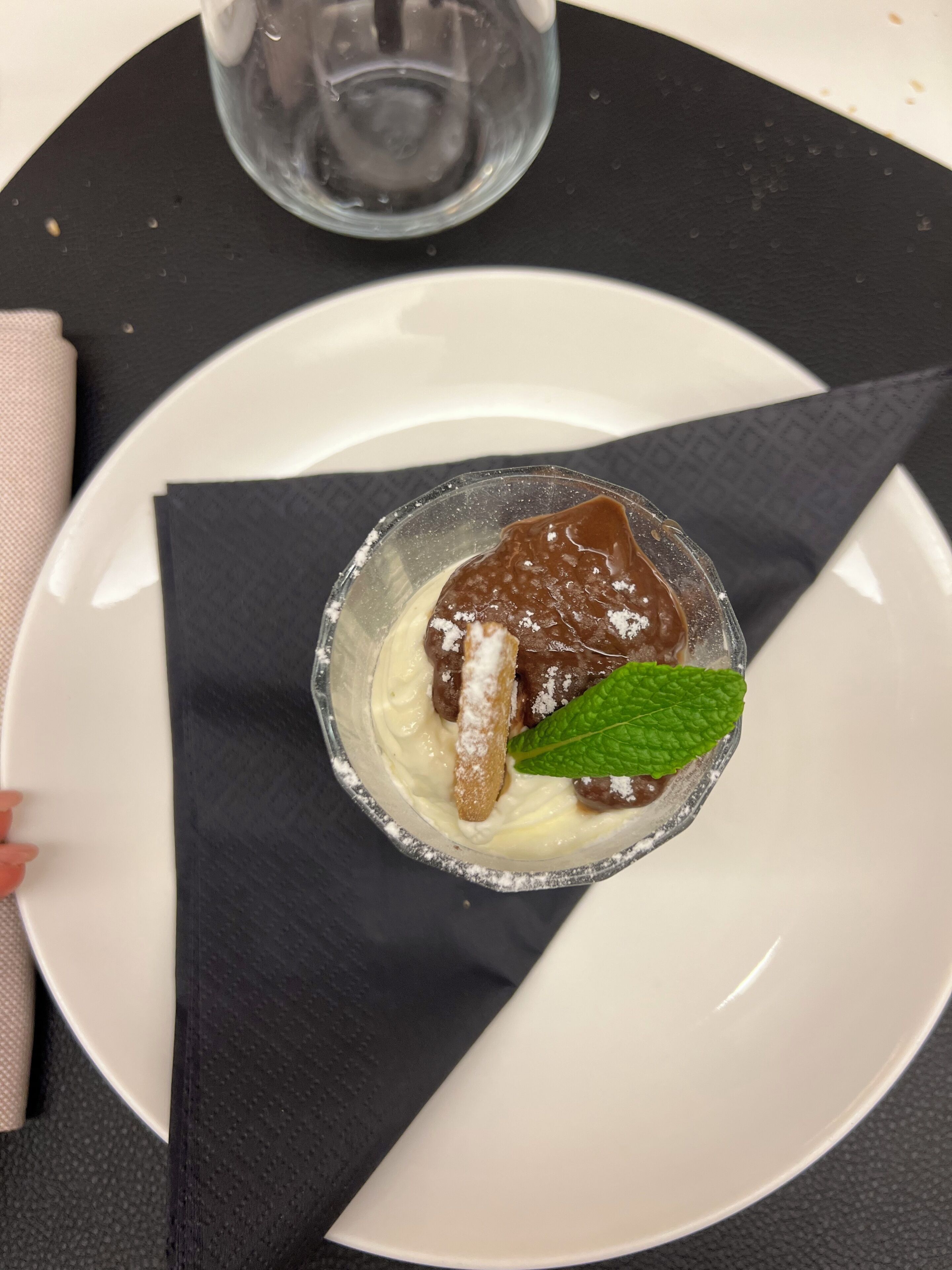 Sobremesa em copo de vidro transparente sobre prato branco, decorada com folha de hortelã e açúcar em pó.