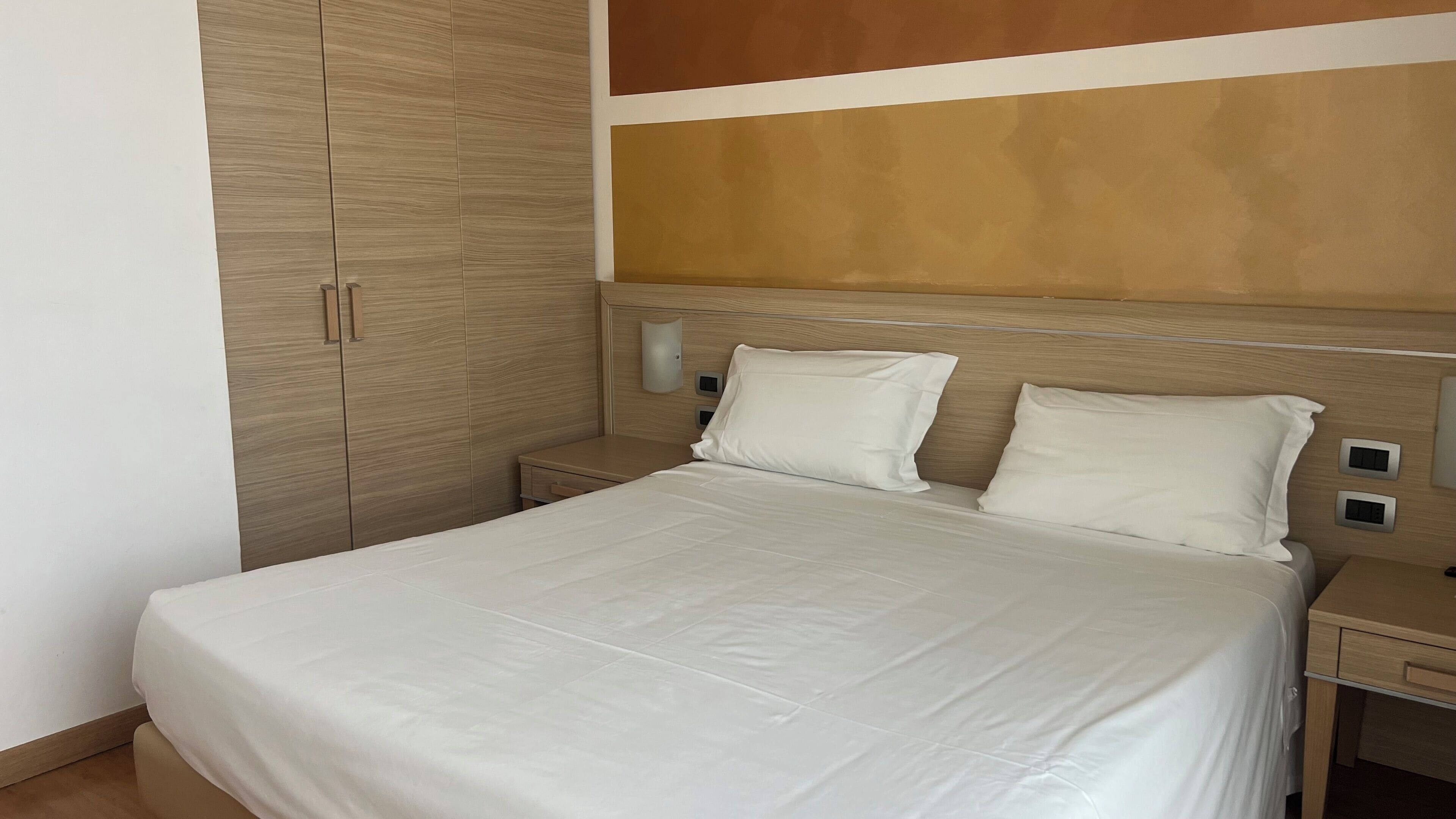Una cama bien hecha con sábanas blancas, un cabecero de madera y una mesita de noche con una lámpara.