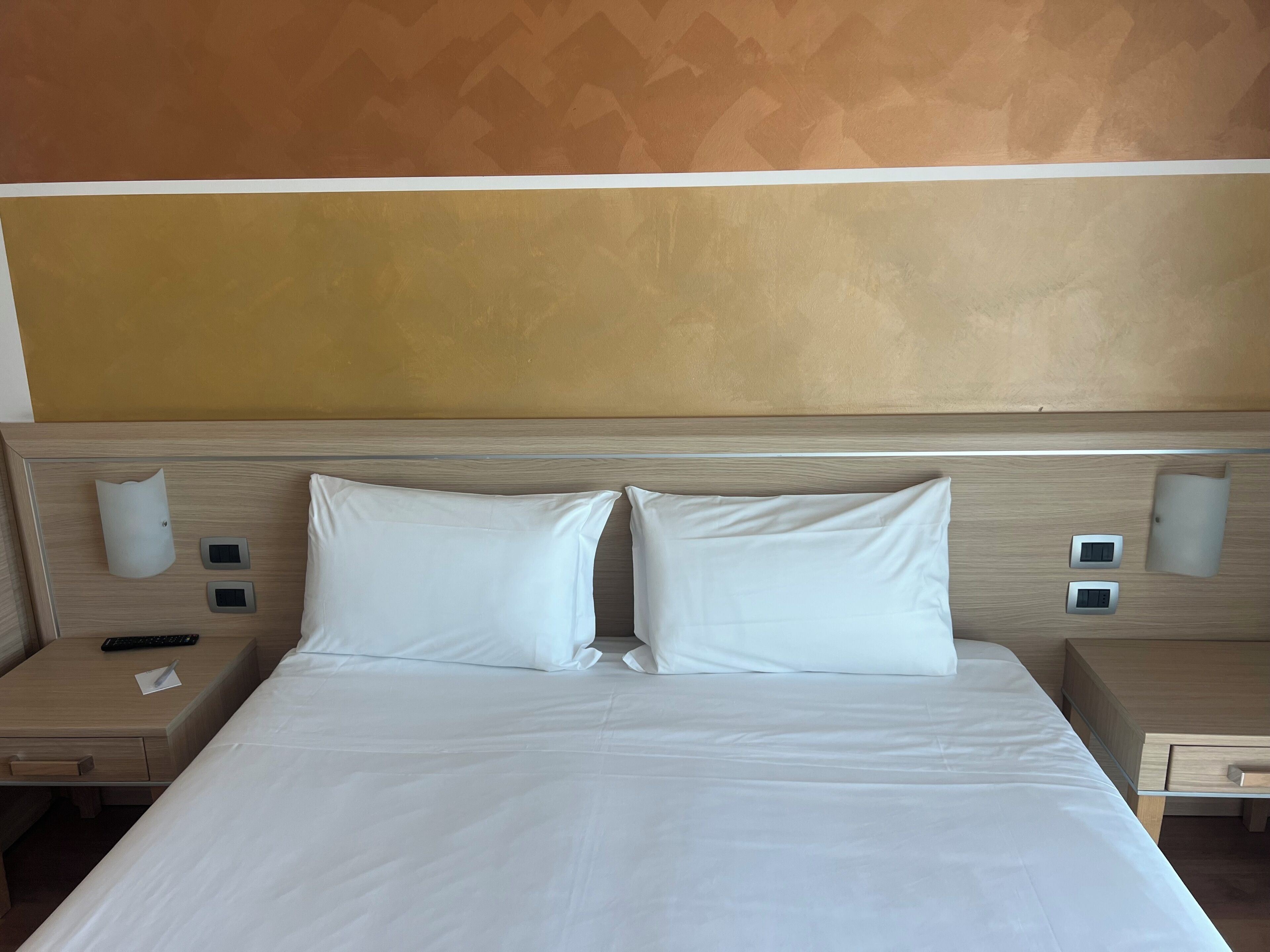 Una habitación de hotel con una cama grande, dos mesitas de noche, un rabillo, un control remoto y una lámpara fijada a la pared.