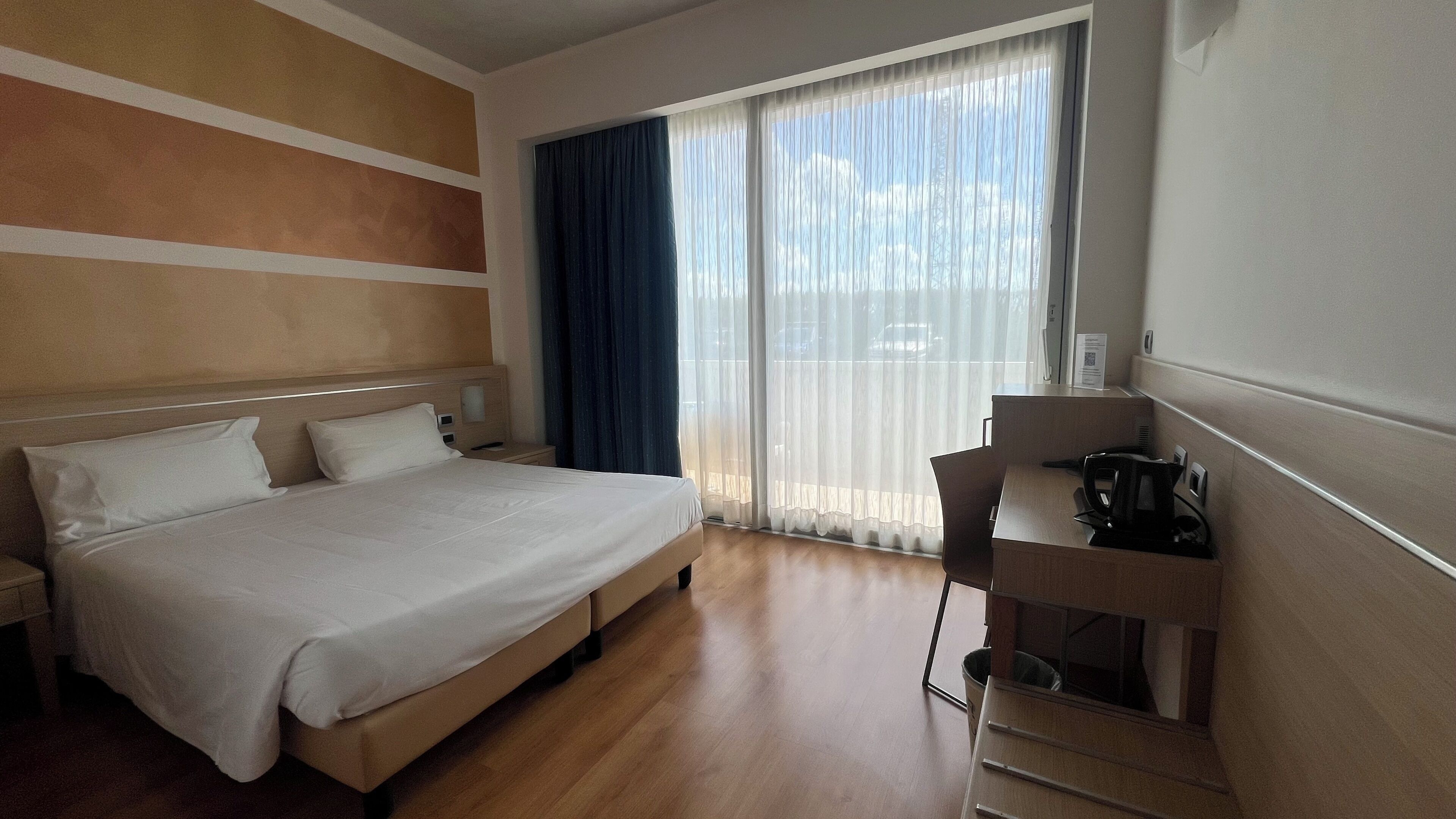 Quarto de hotel com cama, escrivaninha e vista pela janela.