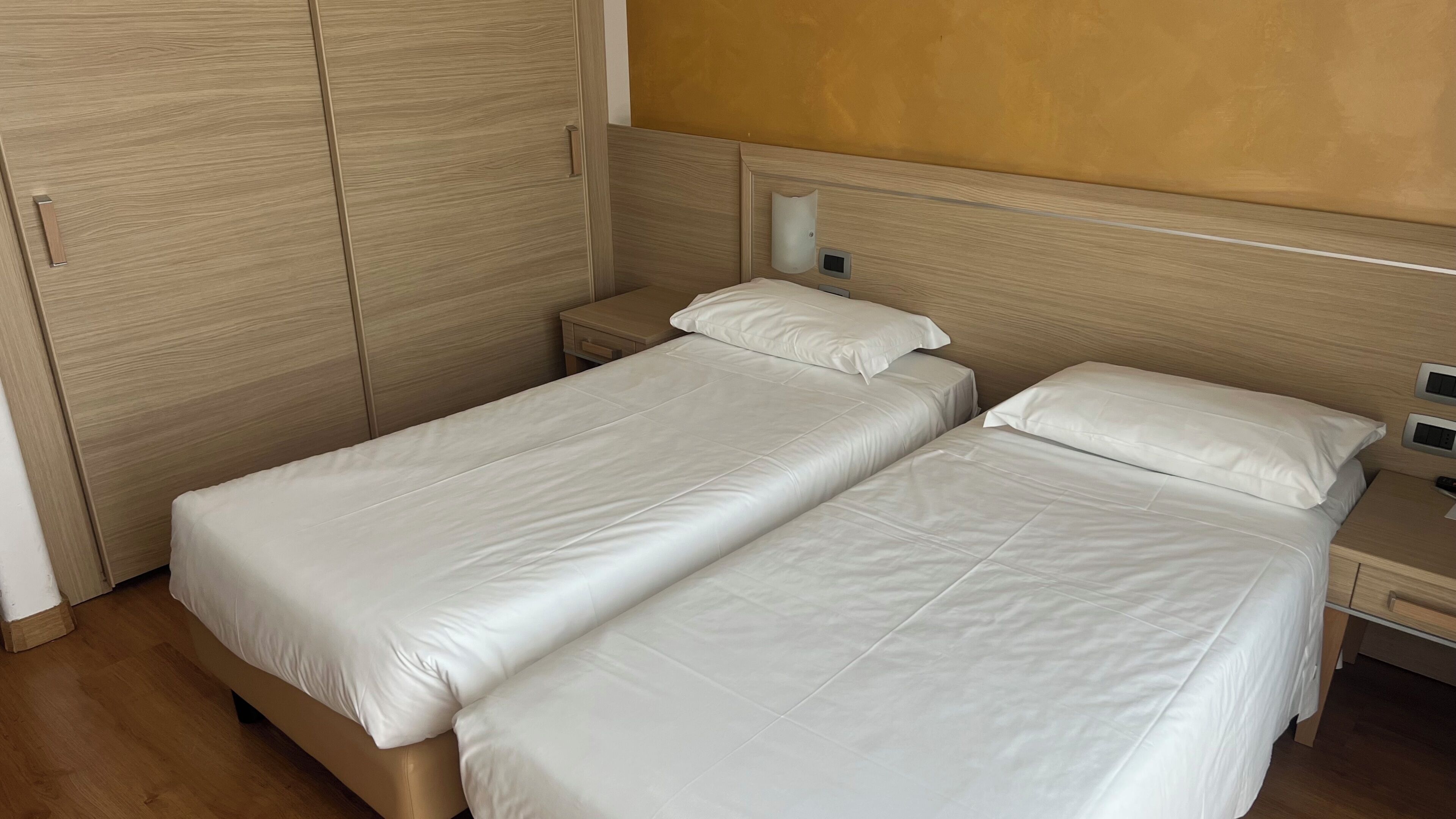 A bedroom with two single beds, wooden wardrobe, bedside tables with lamps, and a wall-mounted light fixture.