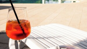 A glass of red drink on a white table by a poolside with lounge chairs and a building in the background. - Hotel Rivus (Peschiera del Garda)