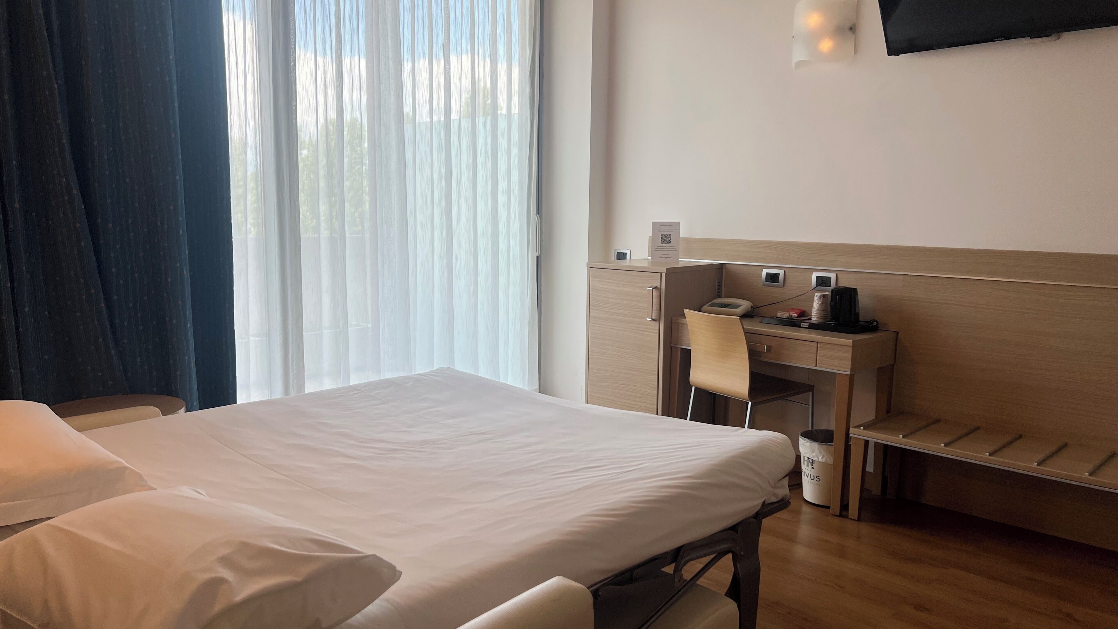 A hotel room with a bed, a desk with a chair, a TV mounted on the wall, and a window with curtains.