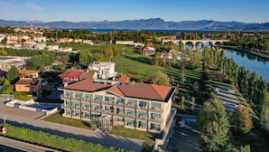A modern hotel with multiple floors, a central courtyard, and a view of a river and mountains in the background. - Hotel Rivus (Peschiera del Garda)
